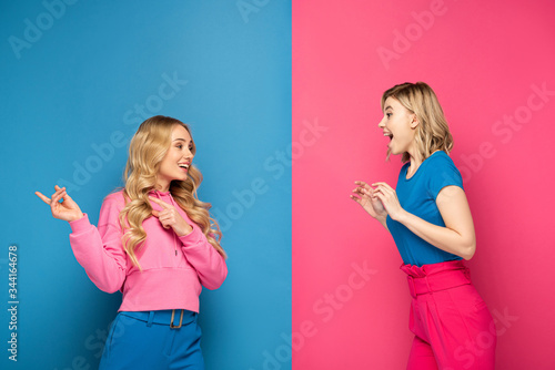 Smiling blonde woman pointing with fingers at shocked sister on pink and blue background