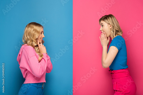 Side view of shocked and smiling blonde sisters looking at each other on pink and blue background
