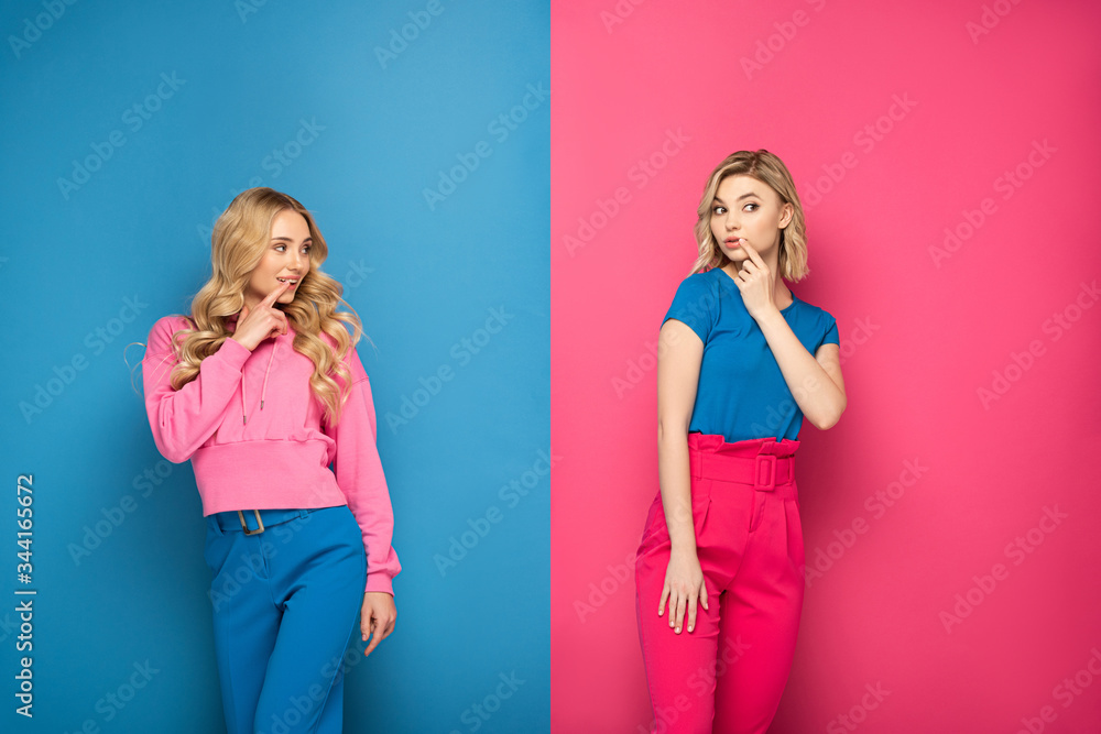 Attractive blonde sisters with fingers near mouths looking away on pink and blue background Attractive blonde sisters with fingers near mouths looking away on pink and blue background