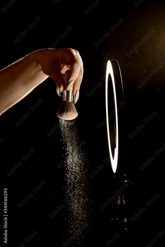 Make-up artist's hand with makeup brushes in front of a ring lamp against a dark background