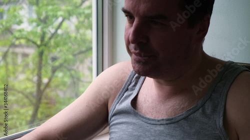 Tired 40 years old man resting, sitting at home near a window, drinking hot tea from a white ceramic mug Quarantine, self isolation, stay at home concept.