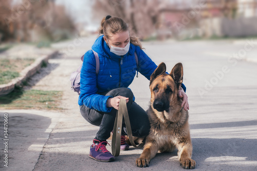 A girl in a protective medical mask walks a dog on the street. Leisure with a pet during quarantine. Walk with a German shepherd in the fresh air. Self-isolation and protection mode.