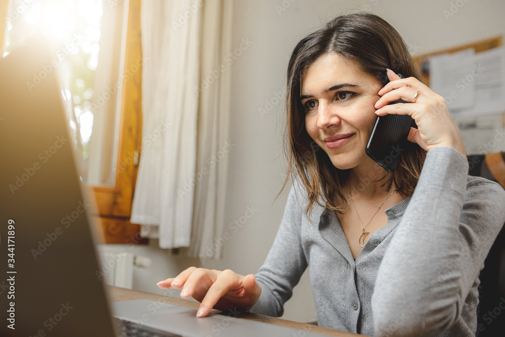 Young woman talking on the phone while working on computer sitting in front a desk next to a window Young woman talking on the phone while working on computer sitting in front a desk next to a window