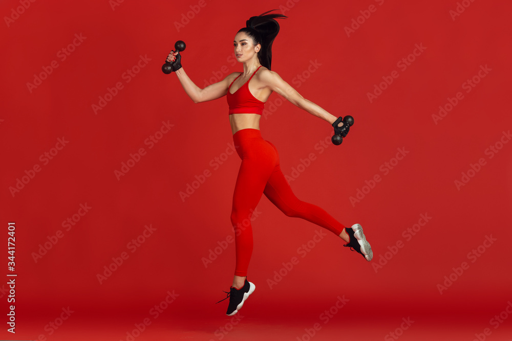 High jump. Beautiful young female athlete practicing in studio, monochrome red portrait. Sportive fit brunette model with weights. Body building, healthy lifestyle, beauty and action concept.