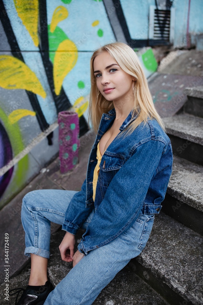 Young blonde woman smiling while sitting on ground against brick wall with graffiti.