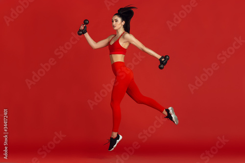 High jump. Beautiful young female athlete practicing in studio, monochrome red portrait. Sportive fit brunette model with weights. Body building, healthy lifestyle, beauty and action concept.
