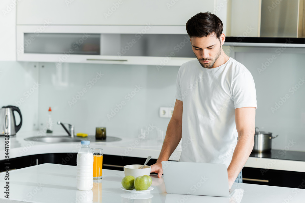 Man using laptop near apples, milk and orange juice on table in kitchen Man using laptop near apples, milk and orange juice on table in kitchen