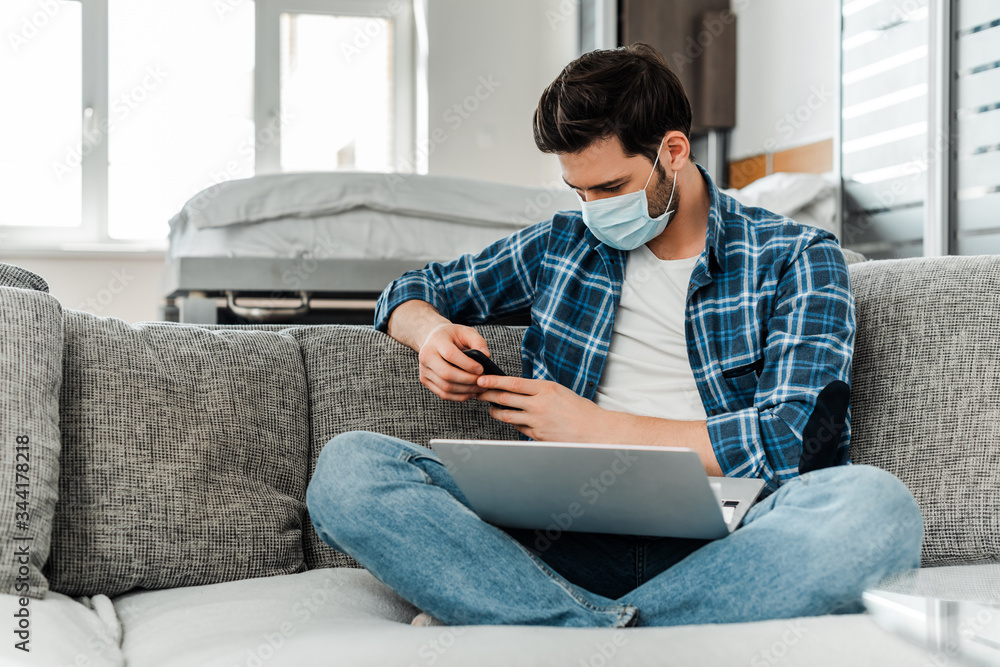 Freelancer in medical mask using smartphone and laptop on couch at home