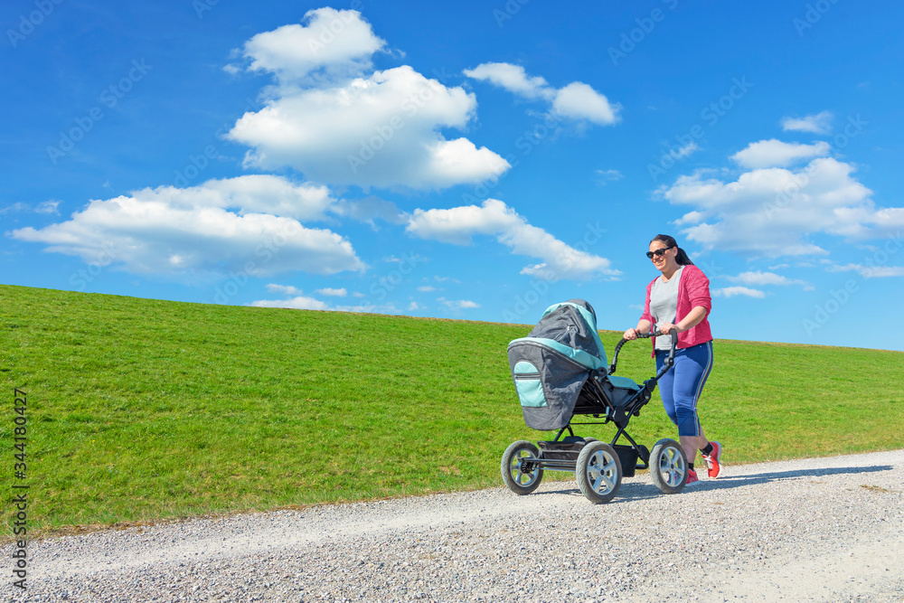 Smiling young mother pushing baby stroller. Green grass and blue sky with clouds background Smiling young mother pushing baby stroller. Green grass and blue sky with clouds background