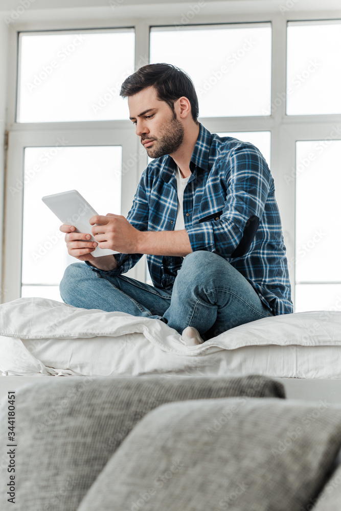 Selective focus of man using digital tablet while sitting on bed near couch at home