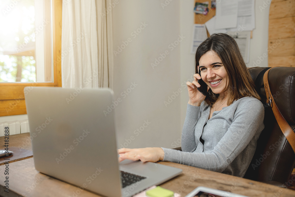 young woman working on laptop and smiling during a phone call. Workplace with wooden desk. young woman working on laptop and smiling during a phone call. Workplace with wooden desk.