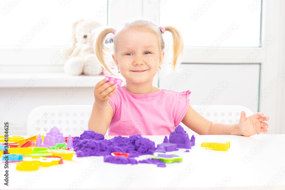 happy Girl plays kinetic sand in quarantine. Blond beautiful girl smiles and plays with purple sand on a white table.