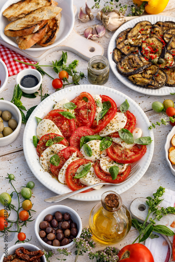 Traditional italian food, Caprese salad made of sliced fresh tomatoes, mozzarella cheese and basil served on a white plate on a wooden table, top view Traditional italian food, Caprese salad made of sliced fresh tomatoes, mozzarella cheese and basil served on a white plate on a wooden table, top view