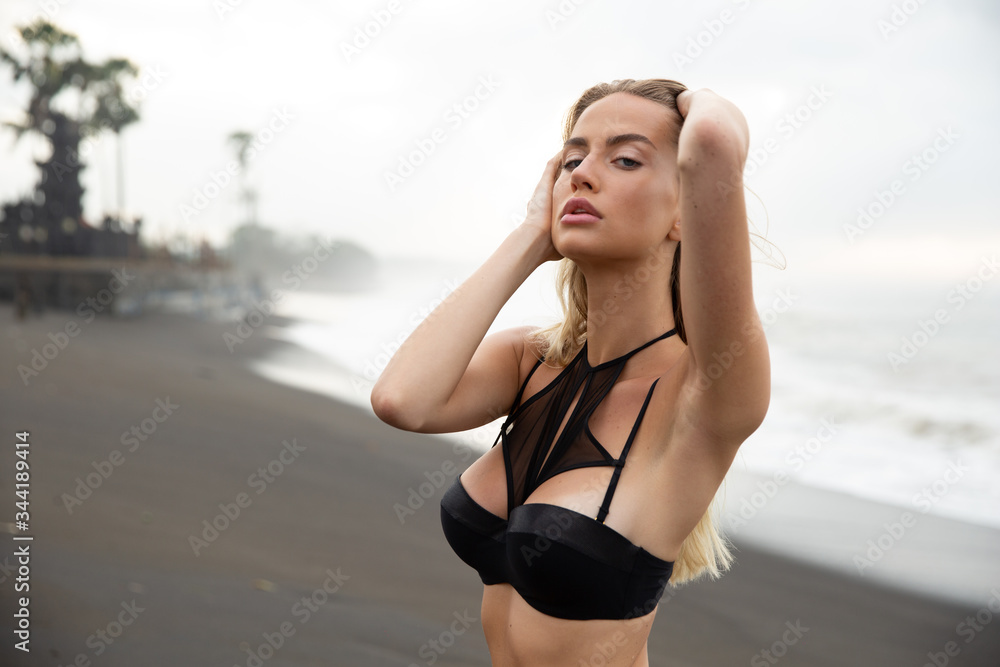 Beautiful sexy girl model lies on black sand beach, in a black swimsuit, with a tanned body, long blonde hair, magnificent breasts and puffy lips. Healthy nature, ocean waves, clouds on Bali island.