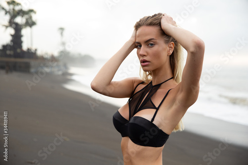 Beautiful sexy girl model lies on black sand beach, in a black swimsuit, with a tanned body, long blonde hair, magnificent breasts and puffy lips. Healthy nature, ocean waves, clouds on Bali island.