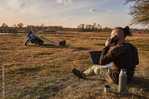 A traveler on a motorcycle talking on the phone working on a laptop admires the sunset