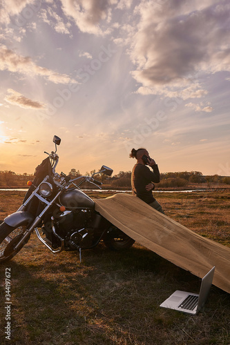 A traveler on a motorcycle talking on the phone working on a laptop admires the sunset