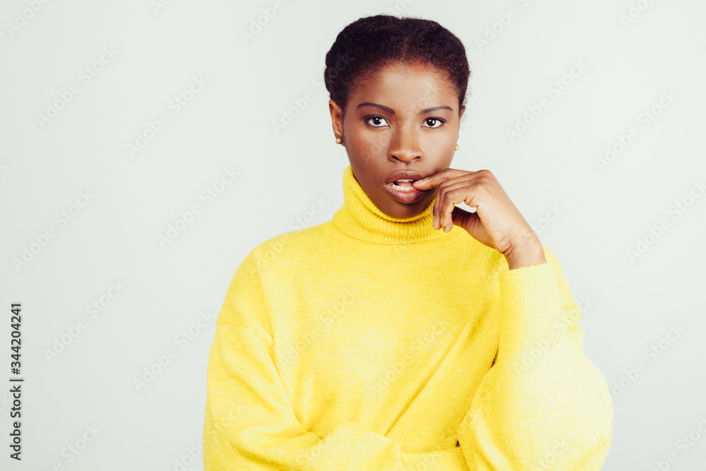 Serious young woman biting finger. Beautiful young African American woman in yellow knitted sweater standing with finger in mouth and looking at camera. Emotion and beauty concept Serious young woman biting finger. Beautiful young African American woman in yellow knitted sweater standing with finger in mouth and looking at camera. Emotion and beauty concept