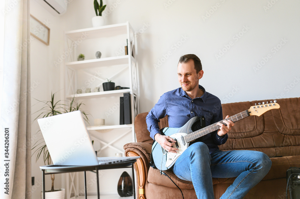 Online studying. A young man is watching video tutorial, video classes how to play guitar, he sits on couch with electric guitar and combo amp near