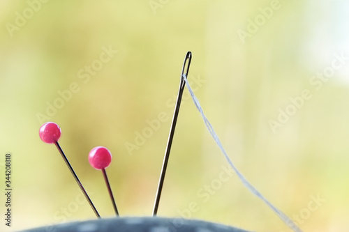 Needle with thread and pink pins on pincushion for handmade sewing cloth with green blurred bokeh background close-up. © Jedsada Naeprai