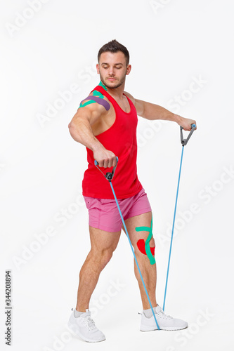 Kinesiology taping. Kinesiology tape on patient knee and shoulder. Young male athlete doing exercises with expender. Post traumatic rehabilitation, sport physical therapy, recovery concept. photo