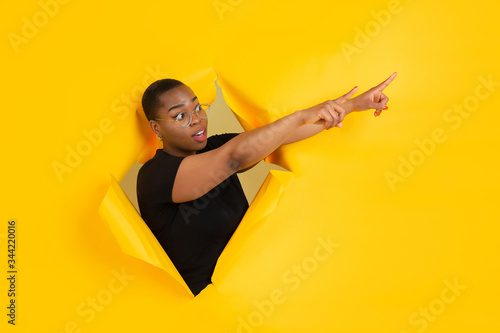 Pointing. Cheerful african-american young woman poses in torn yellow paper background, emotional and expressive. Breaking on, breakthrought. Concept of human emotions, facial expression, sales, ad. © master1305