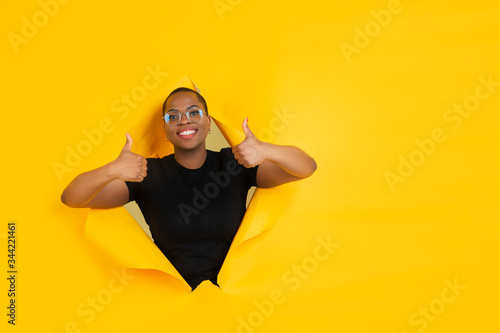 Thumbs up. Cheerful african-american young woman poses in torn yellow paper background, emotional and expressive. Breaking on, breakthrought. Concept of human emotions, facial expression, sales, ad. © master1305
