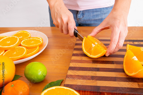 Woman cutting orange with knife on wooden board. White plate with sliced citrus fruits. Fresh mandarin and lime on desk. Cropped side view. Studio shot. Nutrition and vegetarian concept