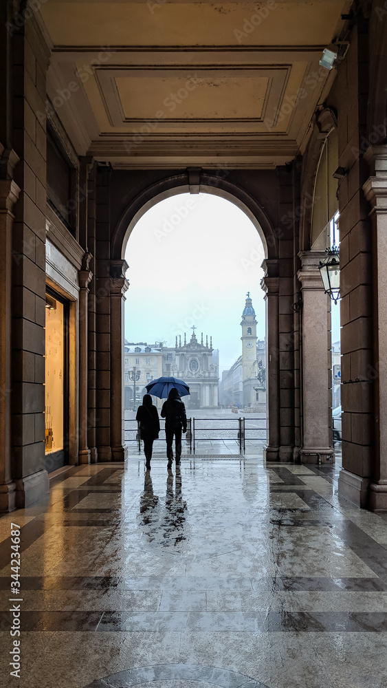 Two people walking with an umbrella in a rainy day under the famous arcades of Turin, Italy, with no other people in sight Two people walking with an umbrella in a rainy day under the famous arcades of Turin, Italy, with no other people in sight