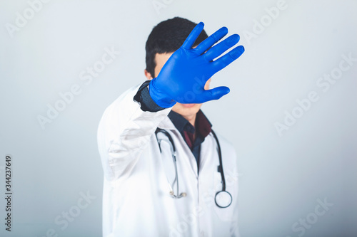 male doctor in a white coat and blue sterile gloves shows a stop gesture with his right hand, gray background