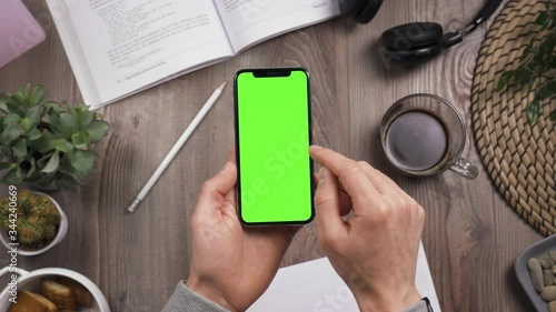 Phone with green screen chroma key in male hands. Swipe left by right hand and forefinger. Top view. Workplace on wooden table with many things on the background. Daylight. POV. 4k video.
