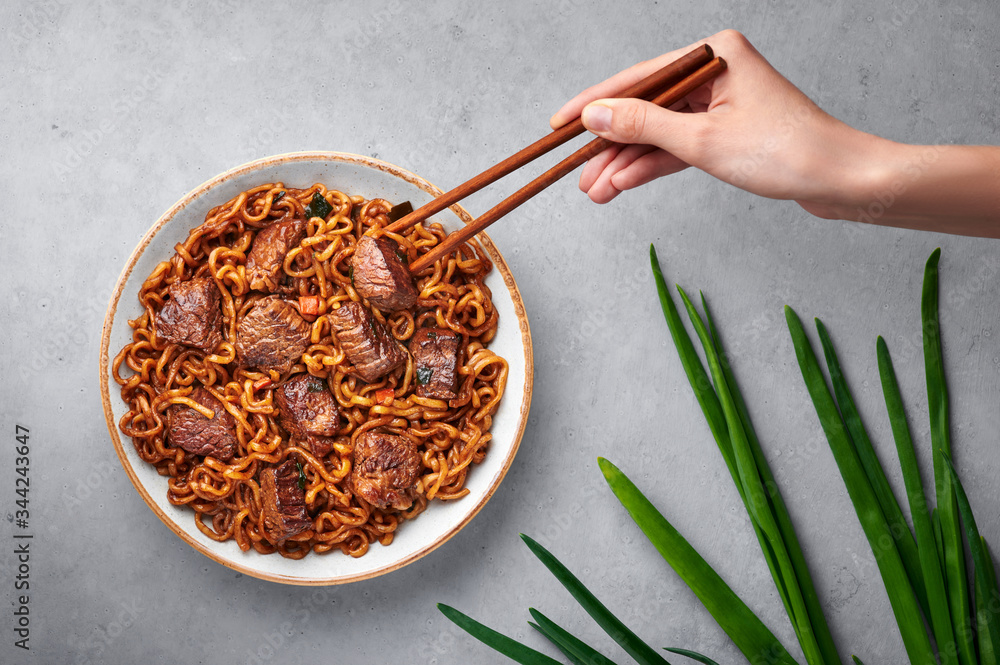 Fototapeta premium Ram-Don or Chapaguri noodles with beef steak in white bowl on grey concrete backdrop. Jjapaguri is a popular south korean dish with ramen and udon noodles and beef steak. Female hand with chopsticks