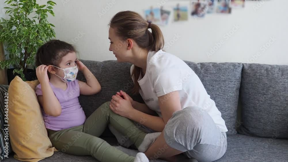 Coronavirus mers. Cute daughter wearing medical mask and her mother helps sitting on sofa at home. Concept of coronavirus COVID-19 pandemic disease symptoms. Home quarantine. Coronavirus mers. Cute daughter wearing medical mask and her mother helps sitting on sofa at home. Concept of coronavirus COVID-19 pandemic disease symptoms. Home quarantine.