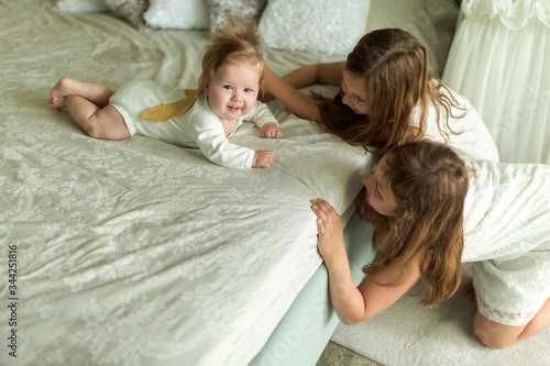 Loving older sisters play with baby brother angel on the bed at home, indoors. Happy loving family. Kids playing in light parents bedroom. Little boy and girls. Love, trust and joy. Childhood.