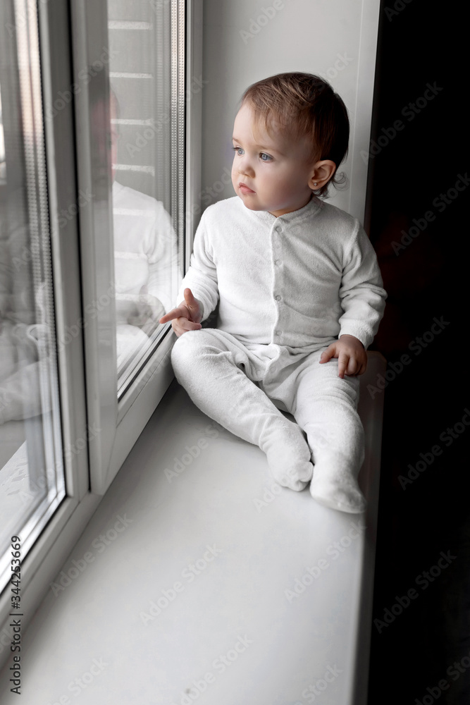 A small child in a white jumpsuit sits on the windowsill of the house and looks out the window. Quarantine and self-isolation A small child in a white jumpsuit sits on the windowsill of the house and looks out the window. Quarantine and self-isolation