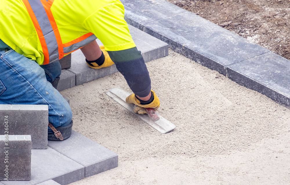 Workman in Safety Jacket Preparing the Foundation of a Patio By Smoothing the Sand with Trowels Workman in Safety Jacket Preparing the Foundation of a Patio By Smoothing the Sand with Trowels