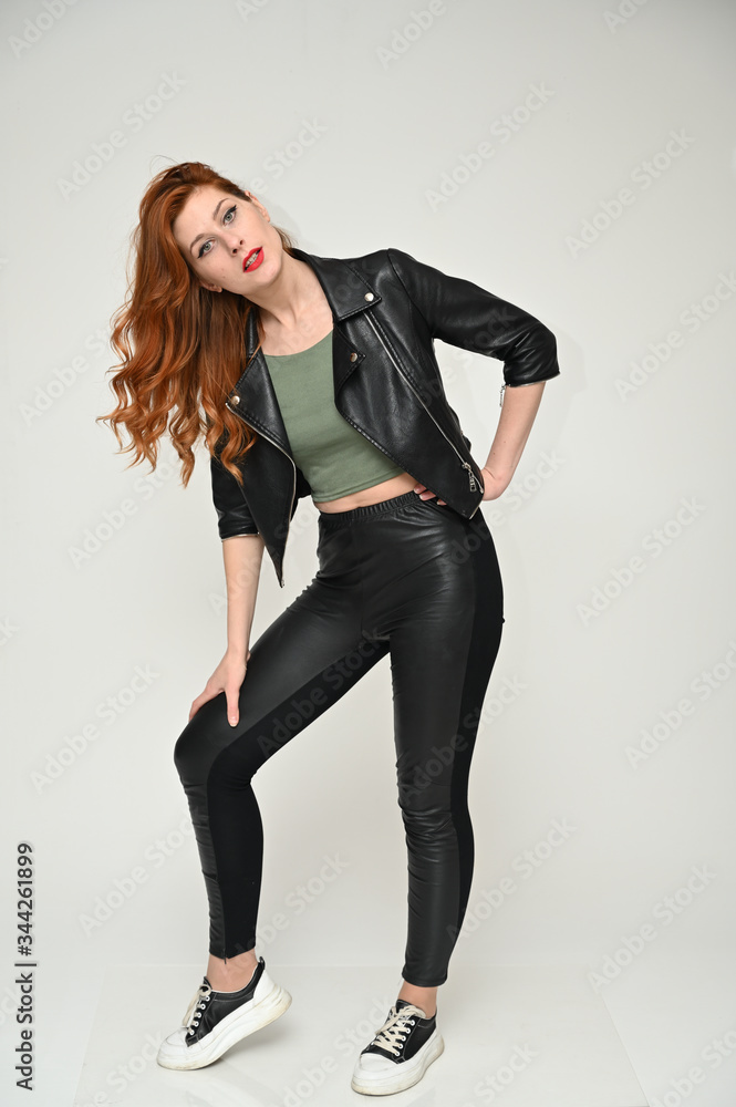 Vertical portrait on a white background of a pretty red-haired young woman. A full-length model with excellent makeup poses in a black jacket and trousers in the studio. Vertical portrait on a white background of a pretty red-haired young woman. A full-length model with excellent makeup poses in a black jacket and trousers in the studio.