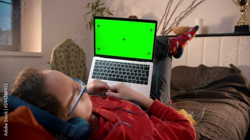 Young man lying on the bed with laptop and eating cheeps - green screen