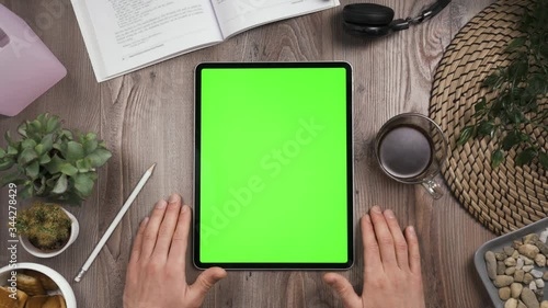 Vertical orientation tab with green chromakey screen. Hand tap on center of the touchscreen then nervously waiting. Top view. Workplace table with many things. Daylight.