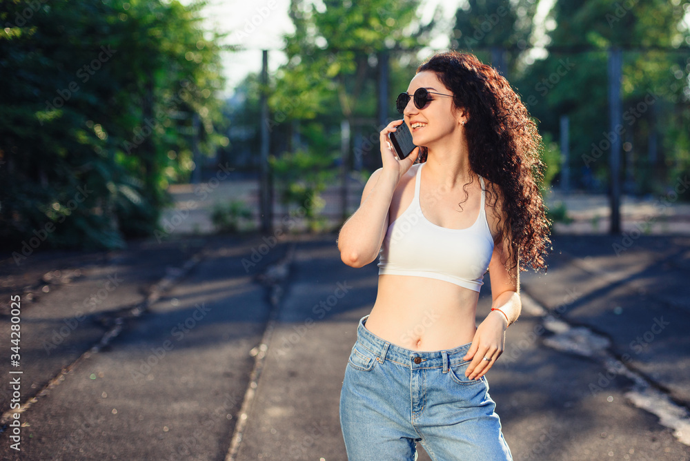 A young brunette in glasses with long curly hair is standing in the street in a white top, jeans are well highlighted. A fun conversation. Green trees in the background A young brunette in glasses with long curly hair is standing in the street in a white top, jeans are well highlighted. A fun conversation. Green trees in the background
