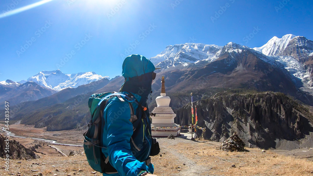 A man hiking next to a stupa with snow caped Annapurna chain in the back, Annapurna Circuit Trek, Himalayas, Nepal. High mountains around. Some prayer's flag next to it. Serenity and calmness.