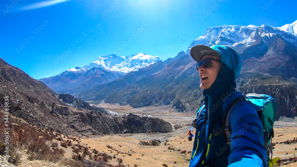 A man hiking and taking a selfie with snow caped Annapurna chain in the back, Annapurna Circuit Trek, Himalayas, Nepal. High mountains around. The man is a bit tired. Serenity and calmness. A man hiking and taking a selfie with snow caped Annapurna chain in the back, Annapurna Circuit Trek, Himalayas, Nepal. High mountains around. The man is a bit tired. Serenity and calmness.