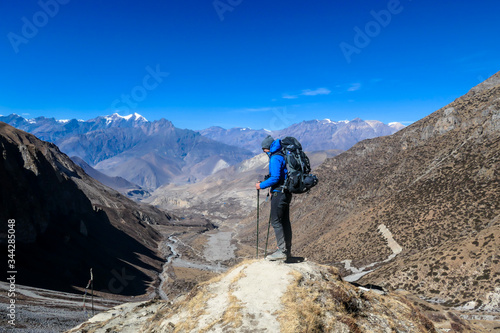 Man hiking through dry path in Himalayan valley, located in Mustang region, Annapurna Circuit Trek in Nepal. He is having a short break, supporting himself on hiking sticks. Harsh landscape. Adventure