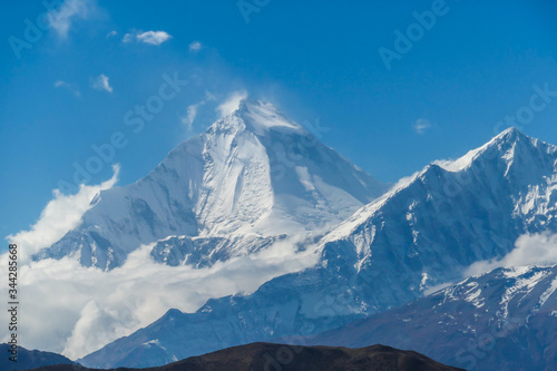A distant view on snow capped Dhaulagiri I, seen from Mustang Valley, Annapurna Circuit Trek in Nepal. Wind blows the snow over the mountain peak. Barren and steep slopes. Harsh landscape. © Chris