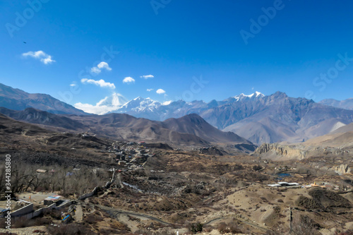 A small Himalayan village, Muktinath, along Annapurna Circuit Trek in Nepal. There is a pathway along a wall. There are only a few buildings. In the back there is high, snow capped Himalayan chain.