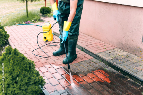 Man cleaning red concrete pavement block using high pressure water cleaner. Paving cleaning concept. Man wearing waders, rubber boots, gloves and waterproof trousers doing spring jobs in the garden photo