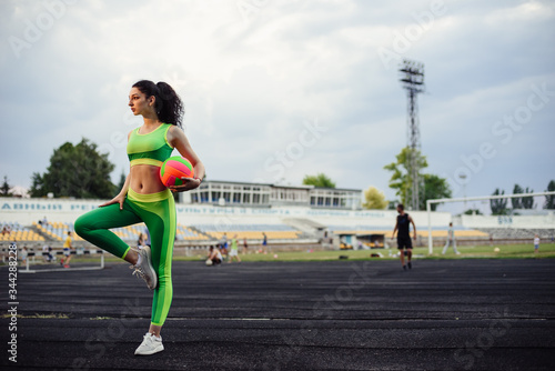 Beautiful curly girl flexing muscles in the stadium. Warm up with the ball. The girl goes in for sports. Light green tracksuit.