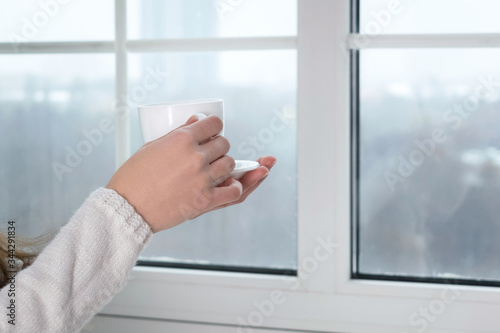 woman's hands holding a cup with hot tea or coffee and looking at the window in her home in the morning evening. Copy space for text, stay home safe at quarantine concept