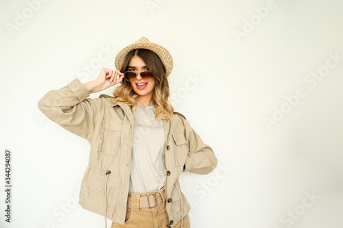 Young beautiful happy woman wearing neutral clothes, sunglasses and a hat on a white background. 