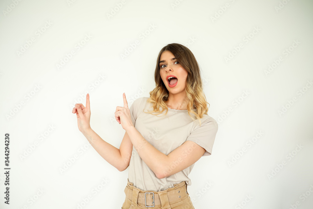 Photo of surprised young woman standing isolated over white wall background. Pointing to copy space, neutral palette. Photo of surprised young woman standing isolated over white wall background. Pointing to copy space, neutral palette.